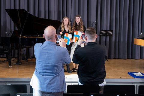 Zwei Absolventinnen stehen mit ihren Urkunden auf der Bühne und werden von ihren Gästen fotografiert. (öffnet Vergrößerung des Bildes)