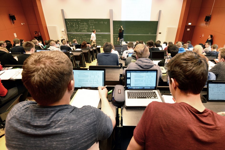 Aufnahme eines Hörsaals mit roten Wänden. Studierende sitzen vor ihren offenen Laptops und hören dem Dozierenden zu.