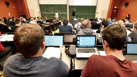 Aufnahme eines Hörsaals mit roten Wänden. Studierende sitzen vor ihren offenen Laptops und hören dem Dozierenden zu.