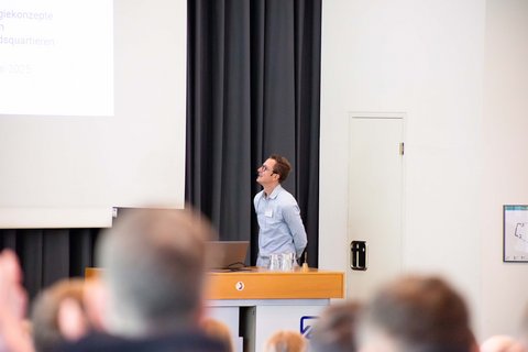 Aufnahme aus der Ferne Richtung Bühne. Ein Speaker steht auf der Bühne und schaut zur Leinwand. (öffnet Vergrößerung des Bildes)