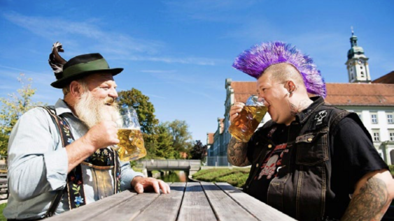 Ein Mann mit weißem Rauschebart und traditioneller süddeutscher Tracht trinkt ein Bier mit einem Punk mit violettem Irokesen und sichtbaren Tätowierungen.
