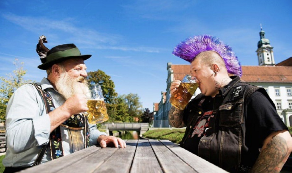 Ein Mann mit weißem Rauschebart und traditioneller süddeutscher Tracht trinkt ein Bier mit einem Punk mit violettem Irokesen und sichtbaren Tätowierungen. 