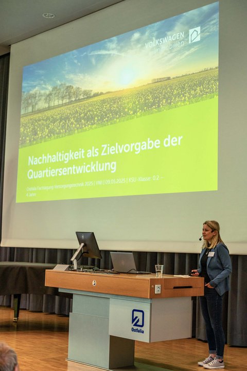 Aufnahme einer Speakerin auf der Bühne in der Aula der Ostfalia Hochschule. Sie schaut in die Richtung der Zuschauer und erzählt über das Thema Nachhaltigkeit als Zielvorgabe für die Quartiersentwicklung. Im Hintergrund ist die passende Präsentation dazu. (öffnet Vergrößerung des Bildes)
