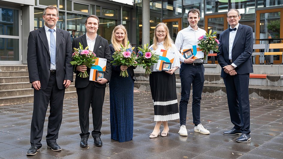 Gruppenbild mit den vier Preisträgerinnen und Preisträgern sowie Dekan und Studiendekan. Die Abgebildeten lächeln in die Kamera. Die Ausgezeichneten halten ihre Awards und Blumensträuße.
