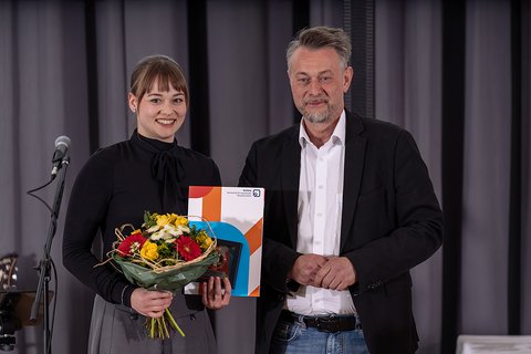 Preisträgerin Anna Riedel in schwarzem Oberteil zusammen mit Bürgermeister Ivica Lukanic in schwarzem Sakko und weißem Hemd. Sie hält ihre Auszeichnung und einen Blumenstrauß. Die Abgebildeten lächeln in die Kamera. (öffnet Vergrößerung des Bildes)