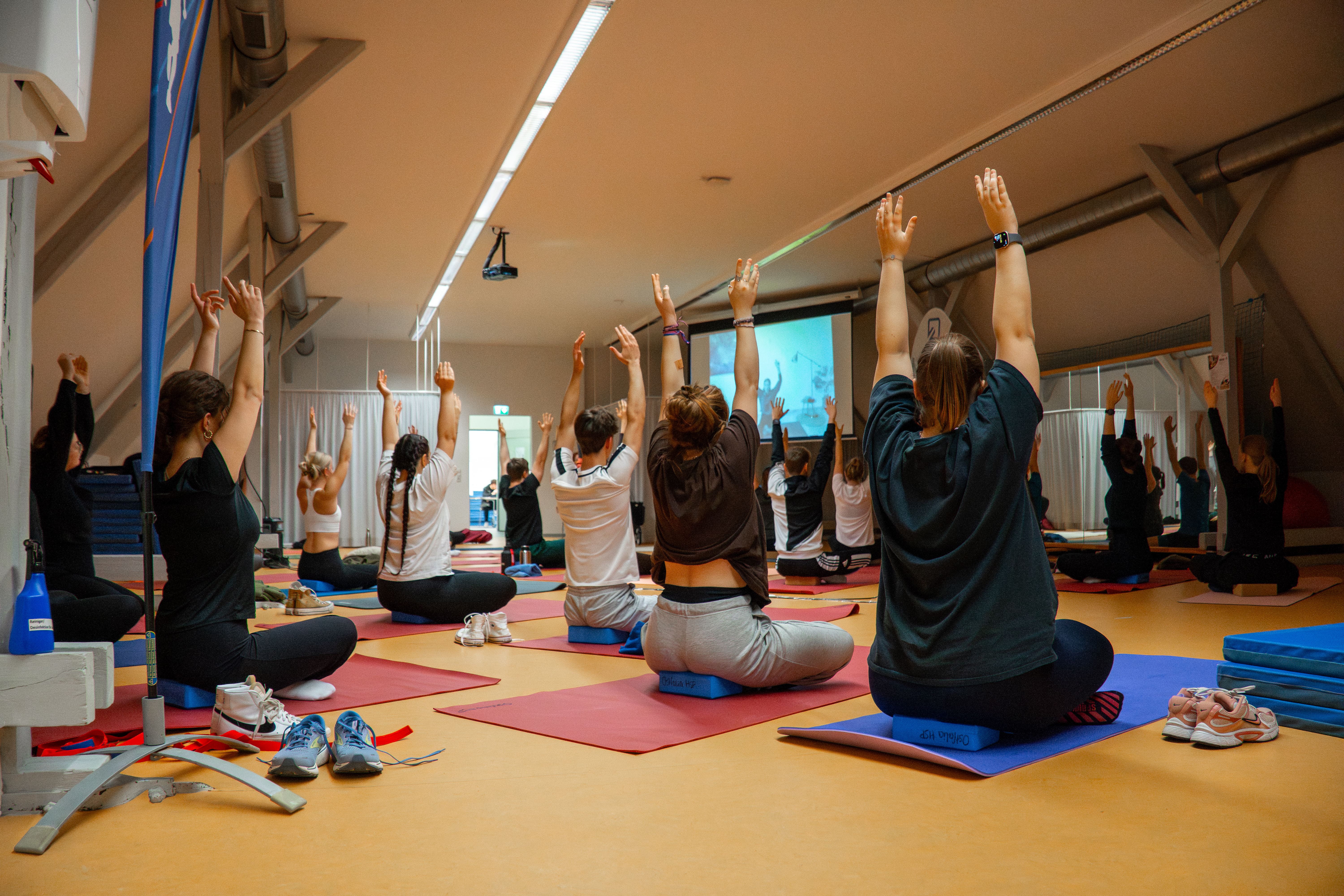Studierende und Mitarbeitende wärmen sich gemeinsam im Yoga im Gymnastikraum der Ostfalia Hochschule in einer Gruppe auf.
