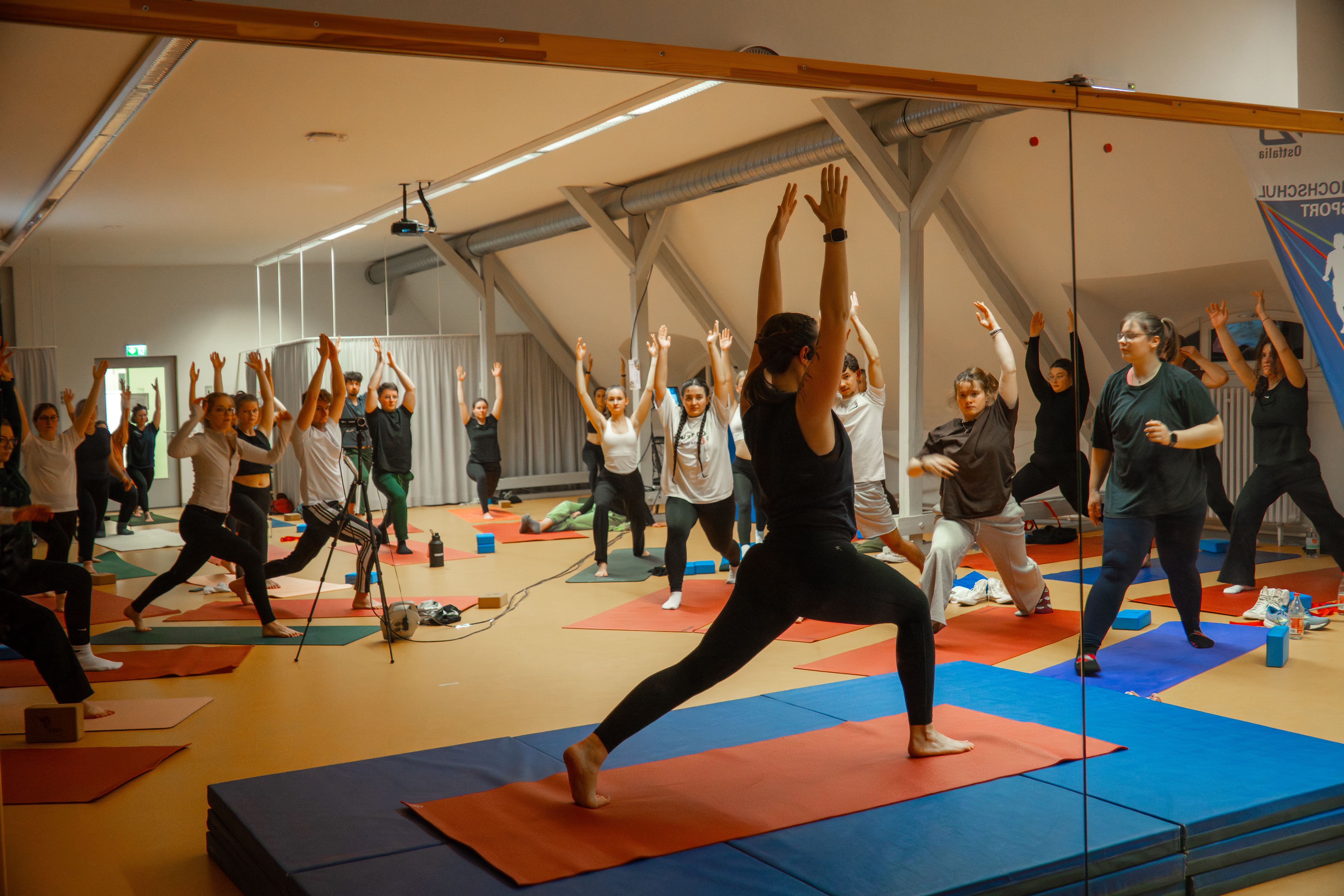 Studierende und Mitarbeitende praktizieren gemeinsam Yoga, normaler Krieger Eins, im Gymnastikraum der Ostfalia Hochschule in einer Gruppe.