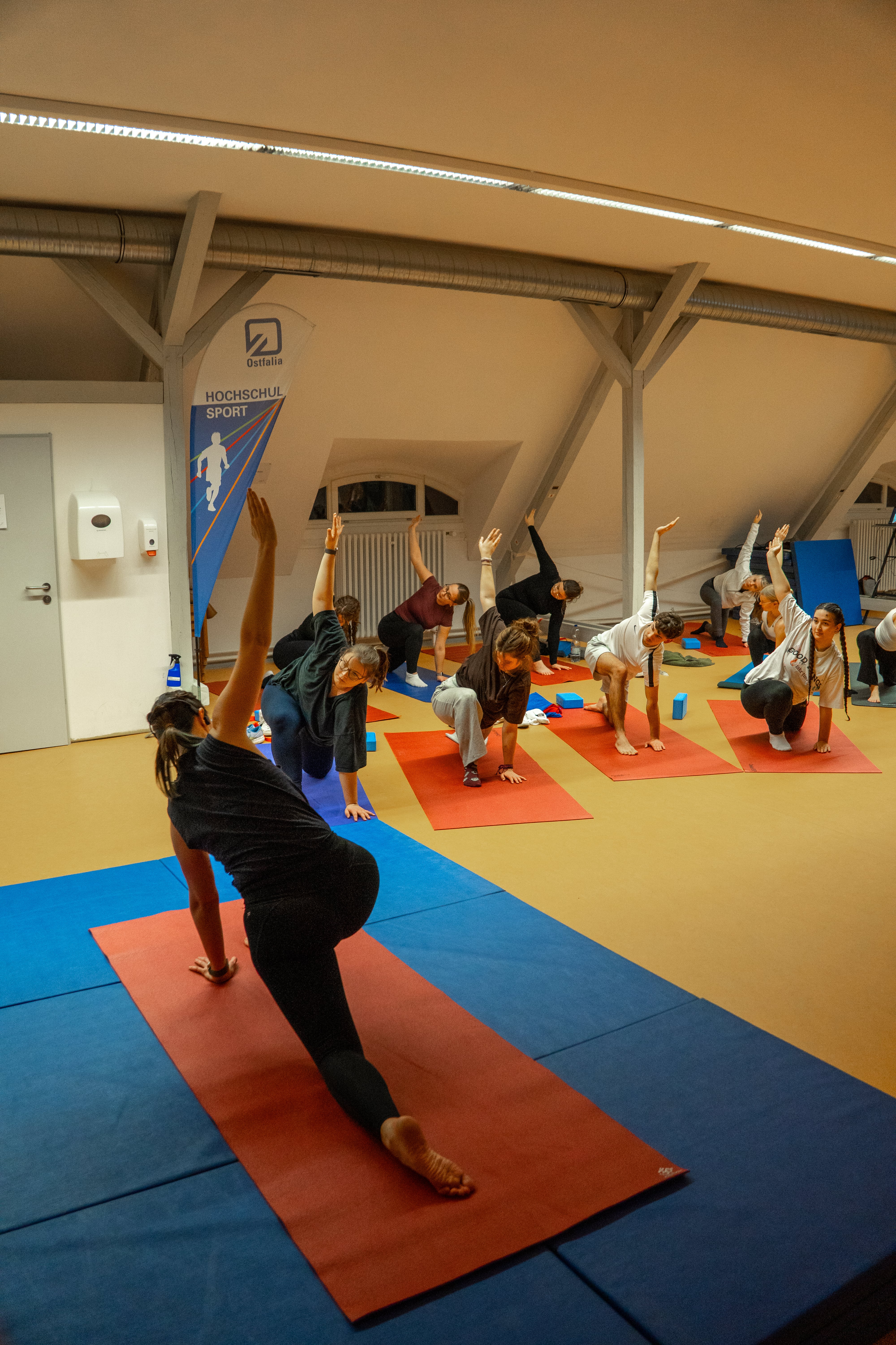 Studierende und Mitarbeitende praktizieren gemeinsam Yoga, Übung geöffneter Krieger eins mit abgelegten Bein, im Gymnastikraum der Ostfalia Hochschule in einer Gruppe.