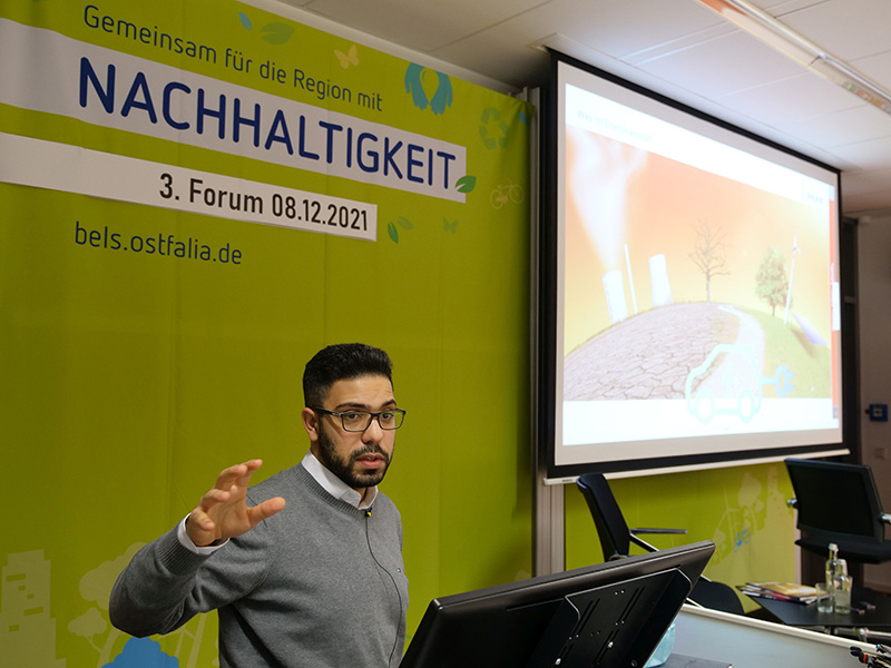 Abbas Seifeddine hält seinen Vortrag beim 3. Forum Nachhaltigkeit. Er steht am Rednerpult, hinter ihm ist eine Leinwand, auf der eine Präsentation gezeigt wird, zu sehen.