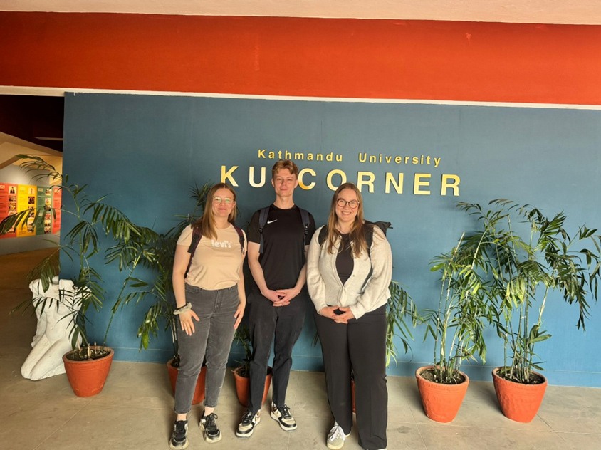 Drei Studierende stehen vor einer blauen Wand auf der in goldenen Buchstaben “Kathmandu University KU CORNER” steht. Die drei stehen vor sechs großen grünen Topfpflanzen und blicken lächelnd in die Kamera.
